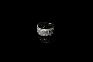 A silver ring encrusted with small sparkling stones is set against a black background, creating a striking contrast. The reflective surface beneath the ring enhances its shine and adds depth to the image.