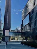 A modern architectural scene featuring a tall industrial chimney and contemporary office buildings with large glass windows. A glass bridge connects the structures, and there is a clear blue sky with few clouds overhead.