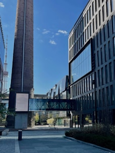 A modern architectural scene featuring a tall industrial chimney and contemporary office buildings with large glass windows. A glass bridge connects the structures, and there is a clear blue sky with few clouds overhead.