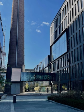 A modern architectural scene featuring a tall industrial chimney and contemporary office buildings with large glass windows. A glass bridge connects the structures, and there is a clear blue sky with few clouds overhead.