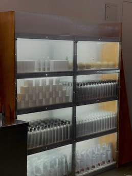A well-lit refrigerated shelf holds an array of products arranged in an organized manner. There are various bottles and containers, possibly different beverages or products, lined up in neat rows. The background and shelves are illuminated with bright white lights, enhancing the visibility of the items. The sides of the shelving unit are made of wood and glass.