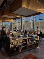 A modern restaurant interior with a central island countertop stocked with dishes and kitchenware. Several people are gathered, some standing while others are seated, chatting and dining. The room is filled with natural light from large windows, and the walls have gold accents. Overhead, stylish lights hang from the ceiling, adding to the contemporary atmosphere.