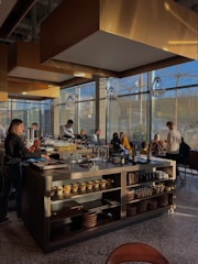 A modern restaurant interior with a central island countertop stocked with dishes and kitchenware. Several people are gathered, some standing while others are seated, chatting and dining. The room is filled with natural light from large windows, and the walls have gold accents. Overhead, stylish lights hang from the ceiling, adding to the contemporary atmosphere.