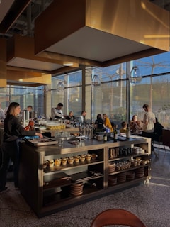 A modern restaurant interior with a central island countertop stocked with dishes and kitchenware. Several people are gathered, some standing while others are seated, chatting and dining. The room is filled with natural light from large windows, and the walls have gold accents. Overhead, stylish lights hang from the ceiling, adding to the contemporary atmosphere.