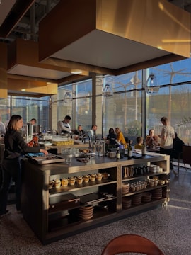 A modern restaurant interior with a central island countertop stocked with dishes and kitchenware. Several people are gathered, some standing while others are seated, chatting and dining. The room is filled with natural light from large windows, and the walls have gold accents. Overhead, stylish lights hang from the ceiling, adding to the contemporary atmosphere.