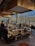 A modern restaurant interior with a central island countertop stocked with dishes and kitchenware. Several people are gathered, some standing while others are seated, chatting and dining. The room is filled with natural light from large windows, and the walls have gold accents. Overhead, stylish lights hang from the ceiling, adding to the contemporary atmosphere.
