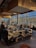 A modern restaurant interior with a central island countertop stocked with dishes and kitchenware. Several people are gathered, some standing while others are seated, chatting and dining. The room is filled with natural light from large windows, and the walls have gold accents. Overhead, stylish lights hang from the ceiling, adding to the contemporary atmosphere.