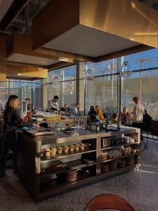 A modern restaurant interior with a central island countertop stocked with dishes and kitchenware. Several people are gathered, some standing while others are seated, chatting and dining. The room is filled with natural light from large windows, and the walls have gold accents. Overhead, stylish lights hang from the ceiling, adding to the contemporary atmosphere.