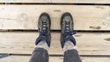 A pair of hiking boots on feet are positioned on wooden planks, showing a top-down perspective. The boots are dark with lighter accents and are worn over thick, light-colored socks and gray pants. The wood surface beneath has visible grain and knots.