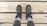 A pair of hiking boots on feet are positioned on wooden planks, showing a top-down perspective. The boots are dark with lighter accents and are worn over thick, light-colored socks and gray pants. The wood surface beneath has visible grain and knots.