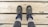 A pair of hiking boots on feet are positioned on wooden planks, showing a top-down perspective. The boots are dark with lighter accents and are worn over thick, light-colored socks and gray pants. The wood surface beneath has visible grain and knots.
