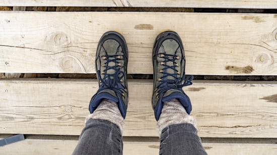A pair of hiking boots on feet are positioned on wooden planks, showing a top-down perspective. The boots are dark with lighter accents and are worn over thick, light-colored socks and gray pants. The wood surface beneath has visible grain and knots.