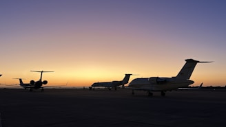 Several private jets are parked on an airport tarmac during a stunning sunset. The sky transitions from a deep blue at the top to a warm orange near the horizon, creating a serene and peaceful atmosphere.
