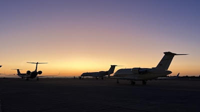 Several private jets are parked on an airport tarmac during a stunning sunset. The sky transitions from a deep blue at the top to a warm orange near the horizon, creating a serene and peaceful atmosphere.