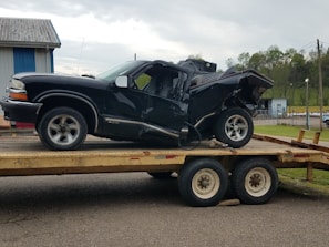 A heavily damaged black vehicle is placed on a trailer, possibly after an accident. The vehicle's rear end is severely crumpled, indicating a significant impact. The scene is outdoors with a building in the background and a cloudy sky overhead.