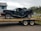 A heavily damaged black vehicle is placed on a trailer, possibly after an accident. The vehicle's rear end is severely crumpled, indicating a significant impact. The scene is outdoors with a building in the background and a cloudy sky overhead.