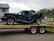 A heavily damaged black vehicle is placed on a trailer, possibly after an accident. The vehicle's rear end is severely crumpled, indicating a significant impact. The scene is outdoors with a building in the background and a cloudy sky overhead.