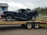 A heavily damaged black vehicle is placed on a trailer, possibly after an accident. The vehicle's rear end is severely crumpled, indicating a significant impact. The scene is outdoors with a building in the background and a cloudy sky overhead.