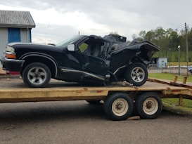A heavily damaged black vehicle is placed on a trailer, possibly after an accident. The vehicle's rear end is severely crumpled, indicating a significant impact. The scene is outdoors with a building in the background and a cloudy sky overhead.