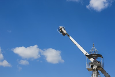 A mechanical amusement park ride is set against a bright blue sky with a few fluffy white clouds. The ride features a crane-like structure with a passenger compartment at the end, and a tower with a viewing platform and an architectural ornament on top.