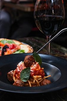 A bowl of spaghetti topped with red tomato sauce, meatballs, and fresh basil is served in a dark bowl. A glass of red wine is placed beside the dish, and a pizza with various toppings is partially visible in the background.