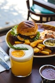 A plate containing a hamburger with a grilled bun and lettuce, accompanied by thick-cut fries and small bowls of dipping sauce. A glass of orange drink garnished with a slice of orange and sprig of rosemary is placed in the foreground. The setting appears to be a cozy dining table with a blurred background.