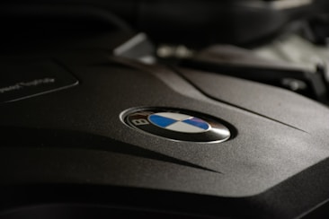 A close-up view of an engine cover with the BMW logo prominently displayed in the center. The surface appears smooth and matte with sharp lines adding a sense of modern design.