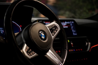 A close-up view of a car's interior featuring a leather-wrapped steering wheel with an emblem. The dashboard is visible with illuminated digital displays and control buttons. Red and blue lighting accents create a modern and sleek appearance.