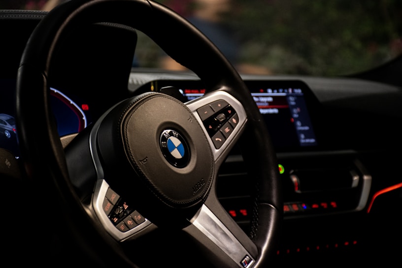 A close-up view of a car's interior featuring a leather-wrapped steering wheel with an emblem. The dashboard is visible with illuminated digital displays and control buttons. Red and blue lighting accents create a modern and sleek appearance.