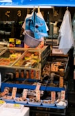 A fish market stall displaying a variety of seafood, including clams and fish, arranged in trays with Japanese labels. The stall is under a blue canopy with hanging bags and a net. There are price tags in yen on display.