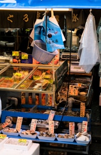 A fish market stall displaying a variety of seafood, including clams and fish, arranged in trays with Japanese labels. The stall is under a blue canopy with hanging bags and a net. There are price tags in yen on display.