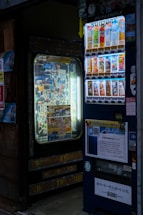 A vending machine situated in a dimly lit area, featuring a variety of drink options displayed brightly. The selection includes colorful cartons and bottles with labels in Japanese. Another machine or display is nearby showing various small items and snacks.