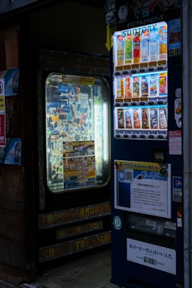A vending machine situated in a dimly lit area, featuring a variety of drink options displayed brightly. The selection includes colorful cartons and bottles with labels in Japanese. Another machine or display is nearby showing various small items and snacks.
