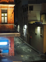 A narrow urban alleyway at night features a corner of a building with a bright blue 'GLOW dental' sign. The street is dimly lit with patches of light from wall-mounted lamps casting shadows on the wet pavement. A blurred figure is walking past, creating a sense of motion. The scene is framed by buildings with different textures and designs, one with red brick and ornate detailing.