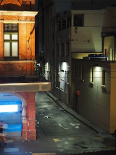 A narrow urban alleyway at night features a corner of a building with a bright blue 'GLOW dental' sign. The street is dimly lit with patches of light from wall-mounted lamps casting shadows on the wet pavement. A blurred figure is walking past, creating a sense of motion. The scene is framed by buildings with different textures and designs, one with red brick and ornate detailing.