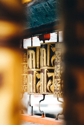 Golden prayer wheels with intricate designs and inscriptions in a traditional style are lined up in a temple setting. The focus is on one prominent wheel, with soft lighting creating a serene atmosphere.