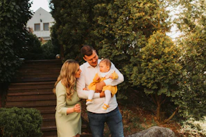 A family is gathered outside near a staircase with lush green trees surrounding them. A man is holding a baby dressed in a bright yellow outfit, while a woman stands nearby in a light green dress, all smiling and interacting with the child.