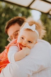 A baby wearing a white headband with a bow rests on the shoulder of an adult, likely a parent. The baby is wearing an orange dress and appears relaxed and comfortable. The background consists of blurred greenery and a structure overhead, suggesting an outdoor setting.