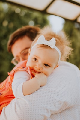 A baby wearing a white headband with a bow rests on the shoulder of an adult, likely a parent. The baby is wearing an orange dress and appears relaxed and comfortable. The background consists of blurred greenery and a structure overhead, suggesting an outdoor setting.