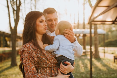 A smiling adoptive couple holding hands, standing in a sunlit park with their young child between them.