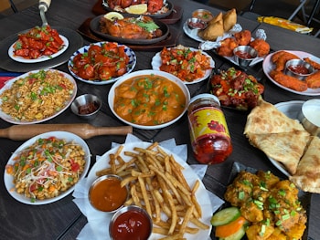 A diverse spread of Indian cuisine on a dark table, including dishes with vibrant colors and various textures. There are plates of bright red chicken dishes, golden fried potatoes with green garnish, and creamy curries. Accompanying these are crispy flatbreads, pickles, samosas, and a selection of dipping sauces. Also visible are rice dishes with vegetables and spices, along with a jar of spicy chili pickles.
