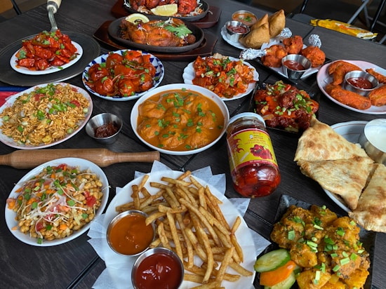 A diverse spread of Indian cuisine on a dark table, including dishes with vibrant colors and various textures. There are plates of bright red chicken dishes, golden fried potatoes with green garnish, and creamy curries. Accompanying these are crispy flatbreads, pickles, samosas, and a selection of dipping sauces. Also visible are rice dishes with vegetables and spices, along with a jar of spicy chili pickles.