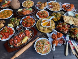 A large assortment of Indian dishes is artfully arranged on a table. The spread includes various curries, shallow-fried breads, grilled meats, fresh salads, and crispy starters. A prominent feature is the vibrant use of spices and colorful garnishes such as fresh herbs and lemon wedges. An array of dips and sauces accompanies the feast.