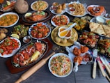 A large assortment of Indian dishes is artfully arranged on a table. The spread includes various curries, shallow-fried breads, grilled meats, fresh salads, and crispy starters. A prominent feature is the vibrant use of spices and colorful garnishes such as fresh herbs and lemon wedges. An array of dips and sauces accompanies the feast.