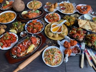 A large assortment of Indian dishes is artfully arranged on a table. The spread includes various curries, shallow-fried breads, grilled meats, fresh salads, and crispy starters. A prominent feature is the vibrant use of spices and colorful garnishes such as fresh herbs and lemon wedges. An array of dips and sauces accompanies the feast.