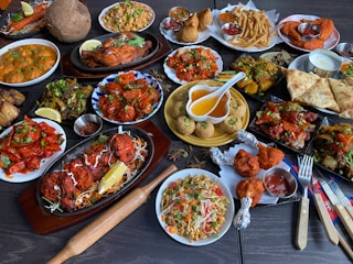 A large assortment of Indian dishes is artfully arranged on a table. The spread includes various curries, shallow-fried breads, grilled meats, fresh salads, and crispy starters. A prominent feature is the vibrant use of spices and colorful garnishes such as fresh herbs and lemon wedges. An array of dips and sauces accompanies the feast.