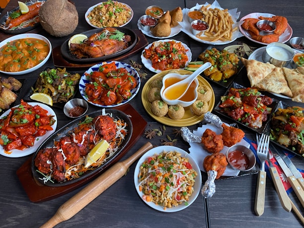 A large assortment of Indian dishes is artfully arranged on a table. The spread includes various curries, shallow-fried breads, grilled meats, fresh salads, and crispy starters. A prominent feature is the vibrant use of spices and colorful garnishes such as fresh herbs and lemon wedges. An array of dips and sauces accompanies the feast.