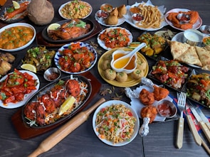 A large assortment of Indian dishes is artfully arranged on a table. The spread includes various curries, shallow-fried breads, grilled meats, fresh salads, and crispy starters. A prominent feature is the vibrant use of spices and colorful garnishes such as fresh herbs and lemon wedges. An array of dips and sauces accompanies the feast.
