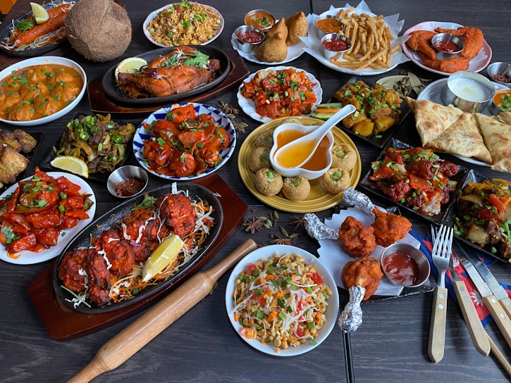A large assortment of Indian dishes is artfully arranged on a table. The spread includes various curries, shallow-fried breads, grilled meats, fresh salads, and crispy starters. A prominent feature is the vibrant use of spices and colorful garnishes such as fresh herbs and lemon wedges. An array of dips and sauces accompanies the feast.