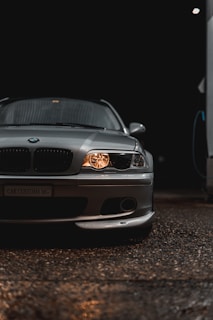 A sleek silver car is prominently displayed, with its headlights on, casting a warm glow against the dark background. The car's front grille and emblem are visible, along with detailed reflections on the smooth surface of the car body. The environment is dimly lit, emphasizing the polished look of the vehicle.